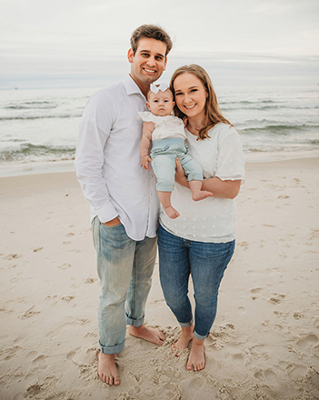 Cameron and wife stand on beach holding their baby girl
